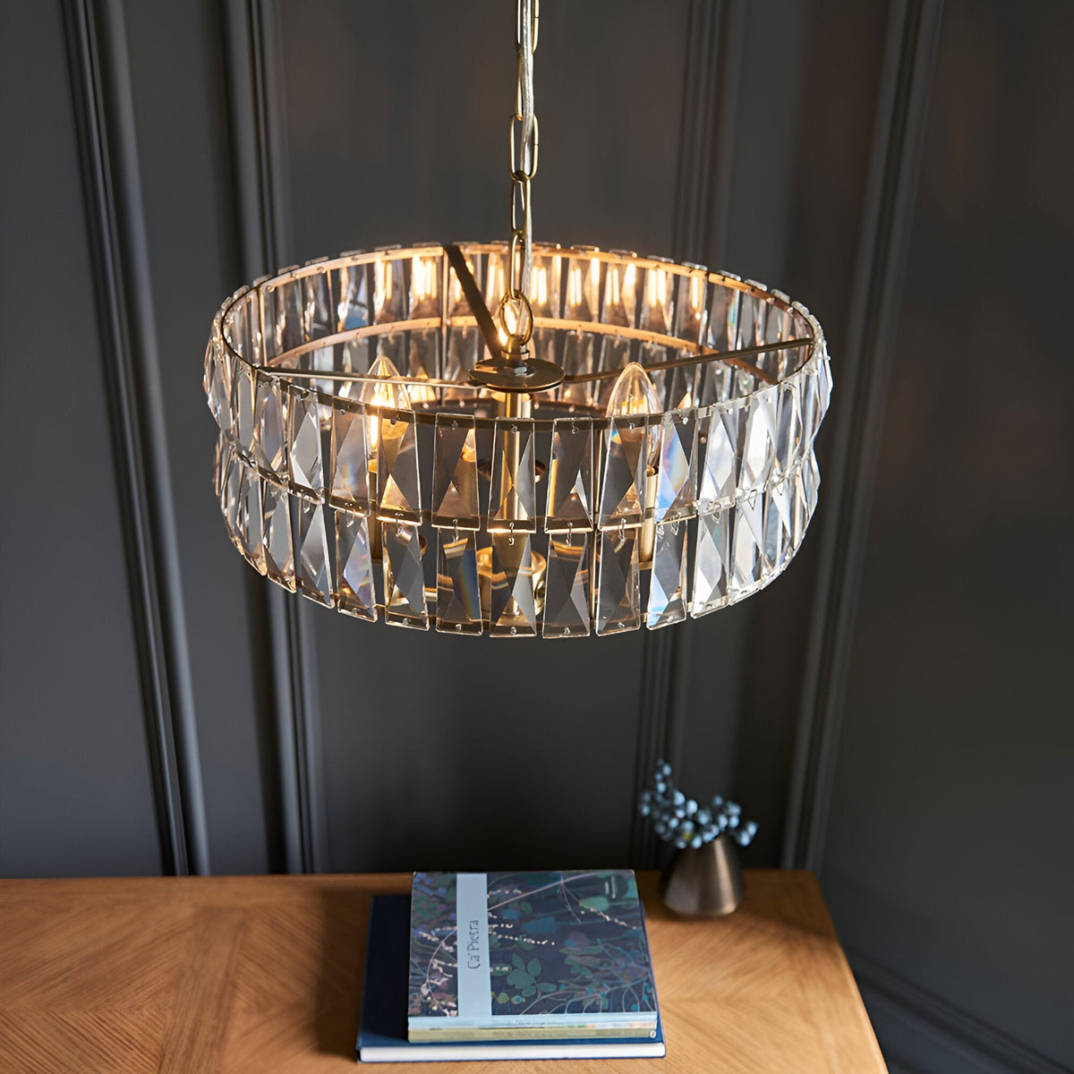 Endon Bevmont 3 light pendant in brass with a drum-shaped tier of cut faceted glass on a chain, shown above a wooden console against a dark panelled wall.