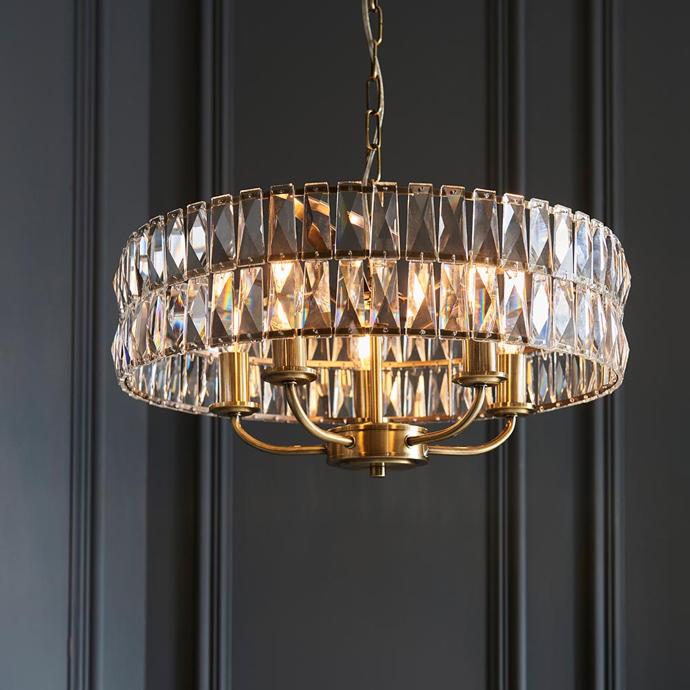 Bevmont 5 Light Pendant in brass with faceted crystal glass drum shade and curved arms, hung on a chain against a dark panelled wall.