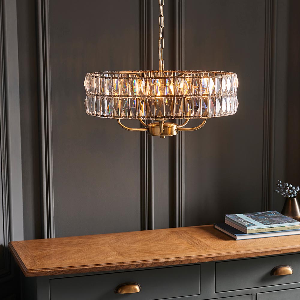 Bevmont 5 Light Pendant in brass with a drum-shaped frame of faceted crystal glass, hung on a chain above a wooden sideboard in a dark panelled room.