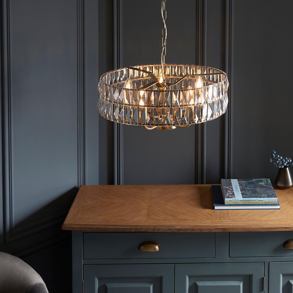 Bevmont 5 Light Pendant in brass with faceted crystal glass and chain suspension, shown lit above a grey sideboard in a dining room setting.