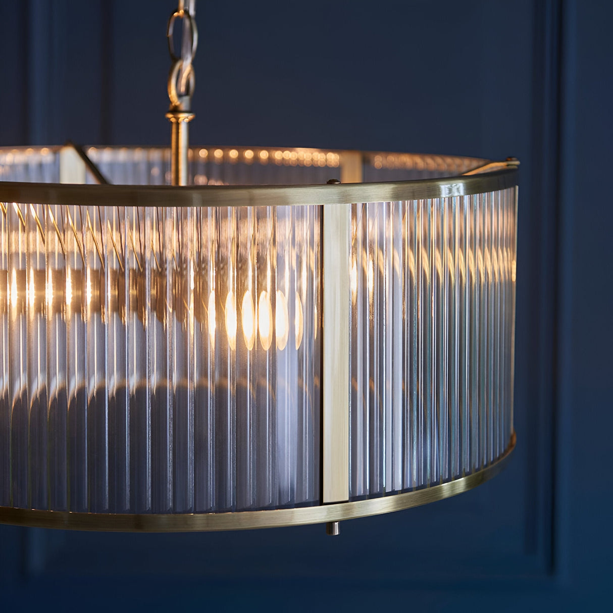 Close-up of the Rothbury 3 Light Pendant in antique brass, showing ribbed glass shade panels and brass frame detail against a dark blue wall.