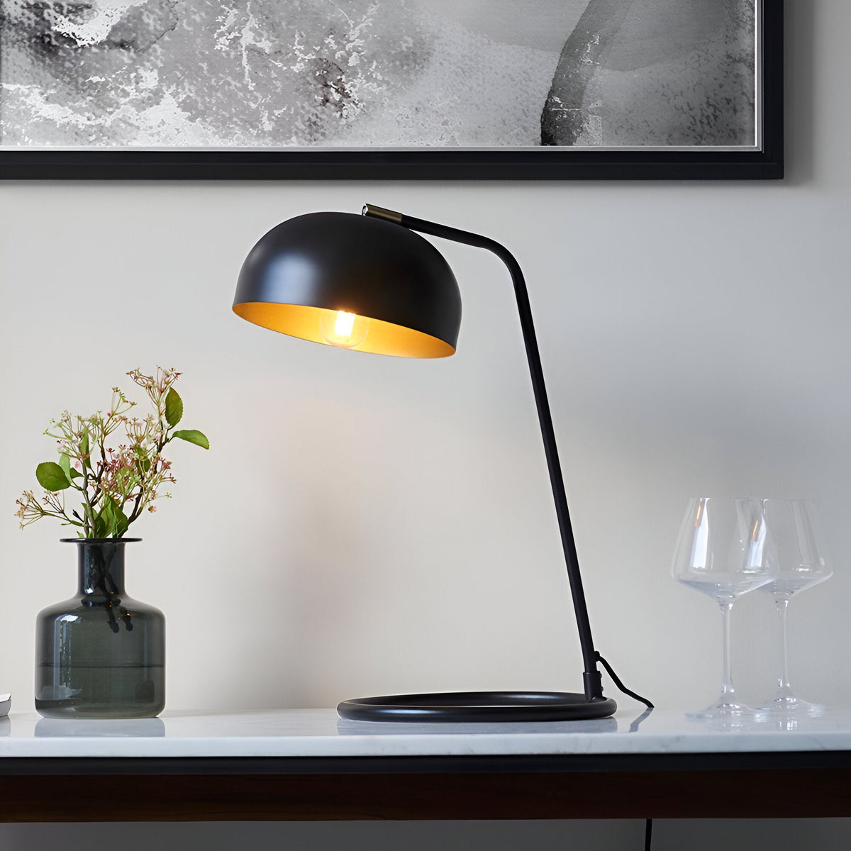 Ashdon Table Lamp in matt black metal with an antique brass inner shade, lit on a marble sideboard beside a vase and glassware.