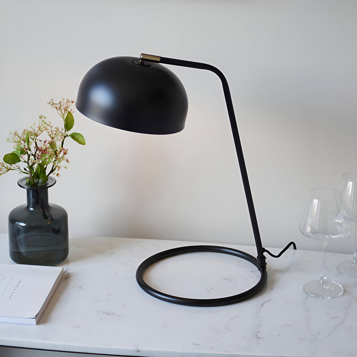 Ashdon Table Lamp in matt black metal with a domed shade, curved stem, and circular base, styled on a marble console beside a vase and glassware.