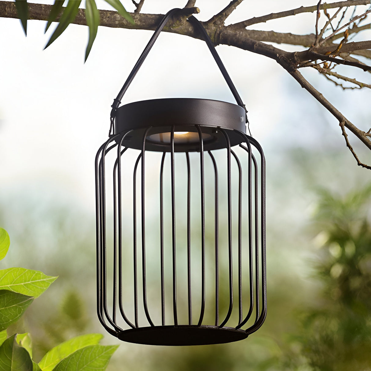 Endon Cranwood LED solar table lamp in matt black, open metal cage design with leather strap handle, hung from a tree branch in a garden setting.