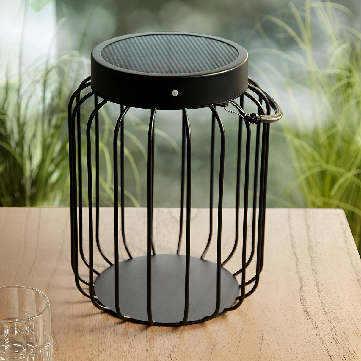 Endon Cranwood LED solar table lamp in matt black with open metal cage frame and hanging handle, shown on a wooden garden table beside grasses.