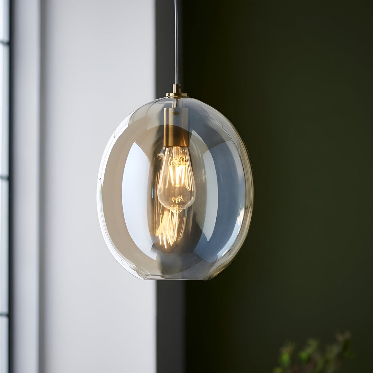 Brockmere Pendant in Brass & Gold, a single-light fixture with an oval champagne lustre glass shade and satin brass fitting, lit against a dark green wall.