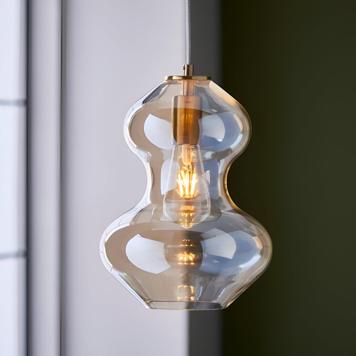 Brockley Pendant in champagne lustre glass with a curved double-bubble shade and satin brass fitting, shown lit with an exposed E27 bulb near a window.