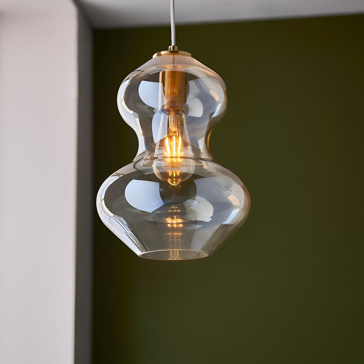 Brockley Pendant in champagne lustre glass with a curved double-bubble shade and satin brass fitting, shown lit against a dark green wall.