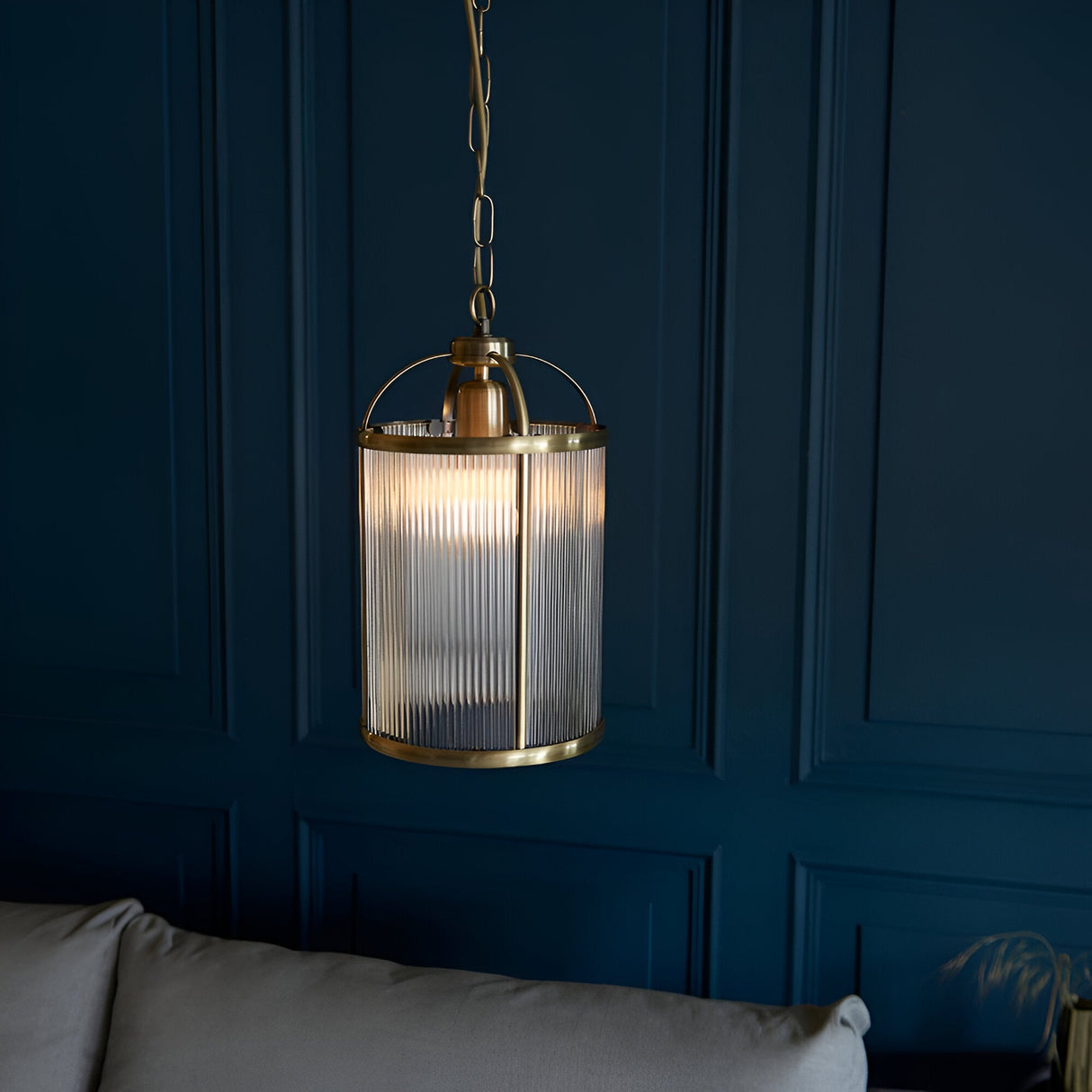 Endon Caldcombe pendant with ribbed glass shade and antique brass frame on chain suspension, shown against a dark blue panelled wall above a grey sofa.