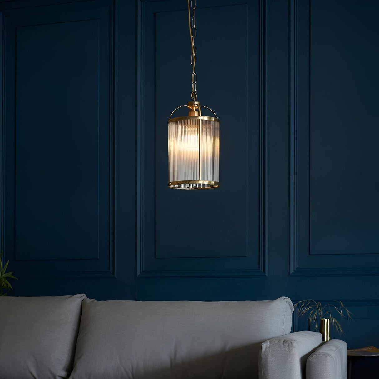 Endon Caldcombe pendant light with ribbed glass shade and antique brass frame on chain, hanging against a dark blue panelled wall above a grey sofa.