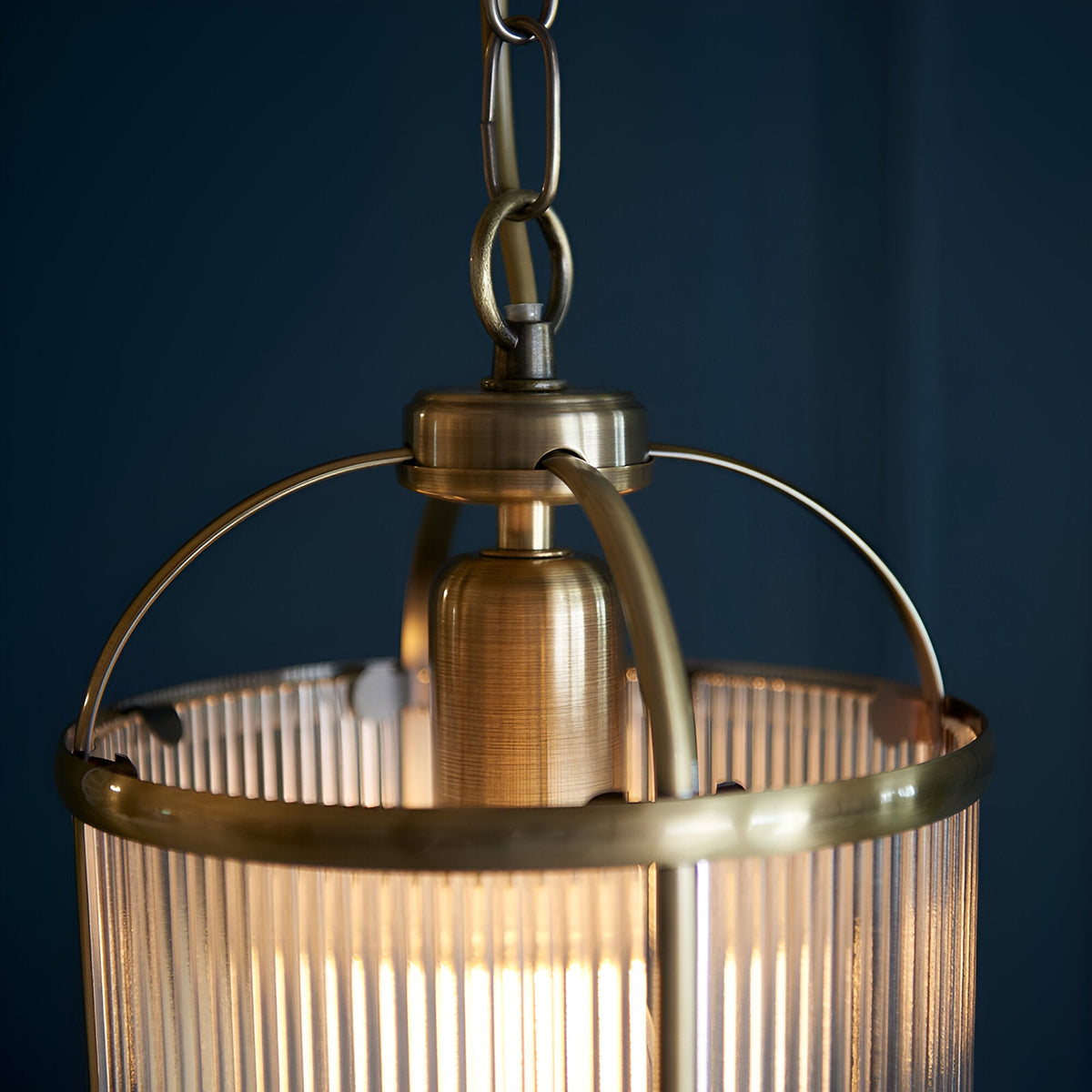Close-up of the Endon Caldcombe pendant showing its antique brass frame, ribbed glass shade, and chain suspension against a dark blue background.