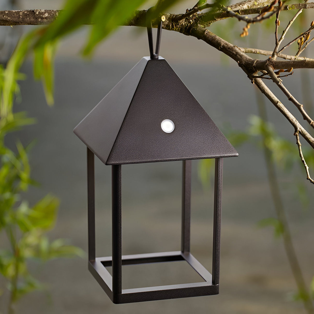 Brenmont Medium Table Lamp in matt black, pyramid-roof lantern design with open frame base, shown hanging outdoors from a tree branch.