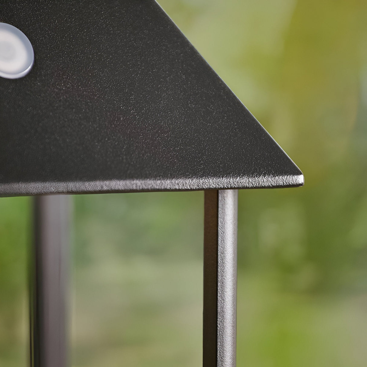 Close-up of the Brenmont Medium Table Lamp's matt black roof edge and slim aluminium support leg, with a blurred green outdoor background.