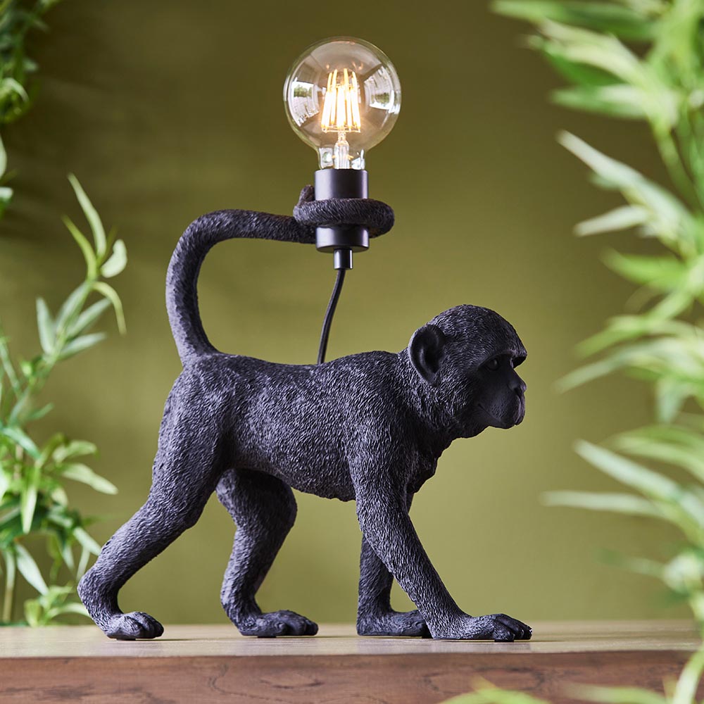 Belvoir Table Lamp in black resin, shaped as a monkey with tail curled around an exposed filament bulb, set against a green foliage backdrop.