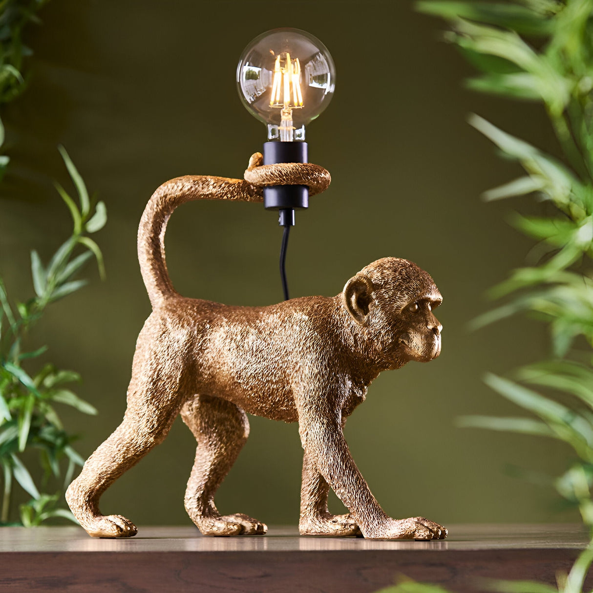 Endon Belvoir table lamp shaped as a gold resin monkey holding an exposed filament bulb above its tail, shown on a shelf with foliage behind.