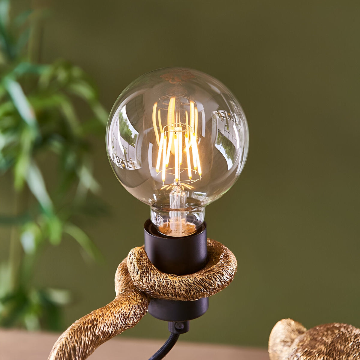 Close-up of the Endon Belvoir table lamp's exposed E27 filament globe bulb held in the gold resin monkey's hand against a green backdrop.