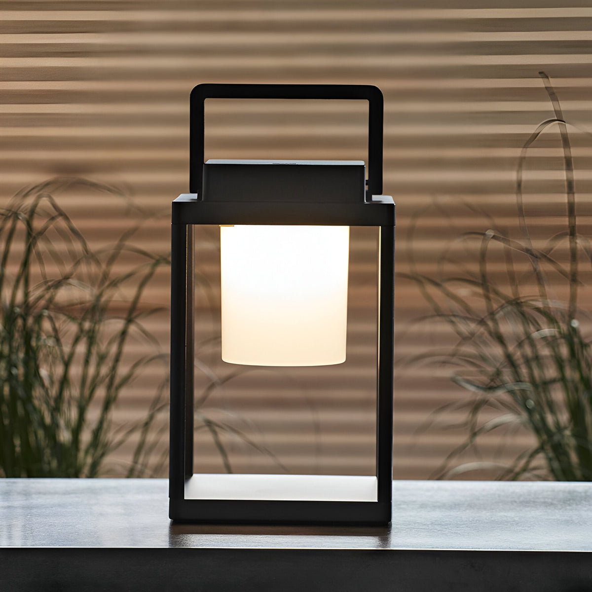 Endon Danedon portable LED table lamp in matt black with white polycarbonate diffuser and carry handle, set on an outdoor table beside grasses.