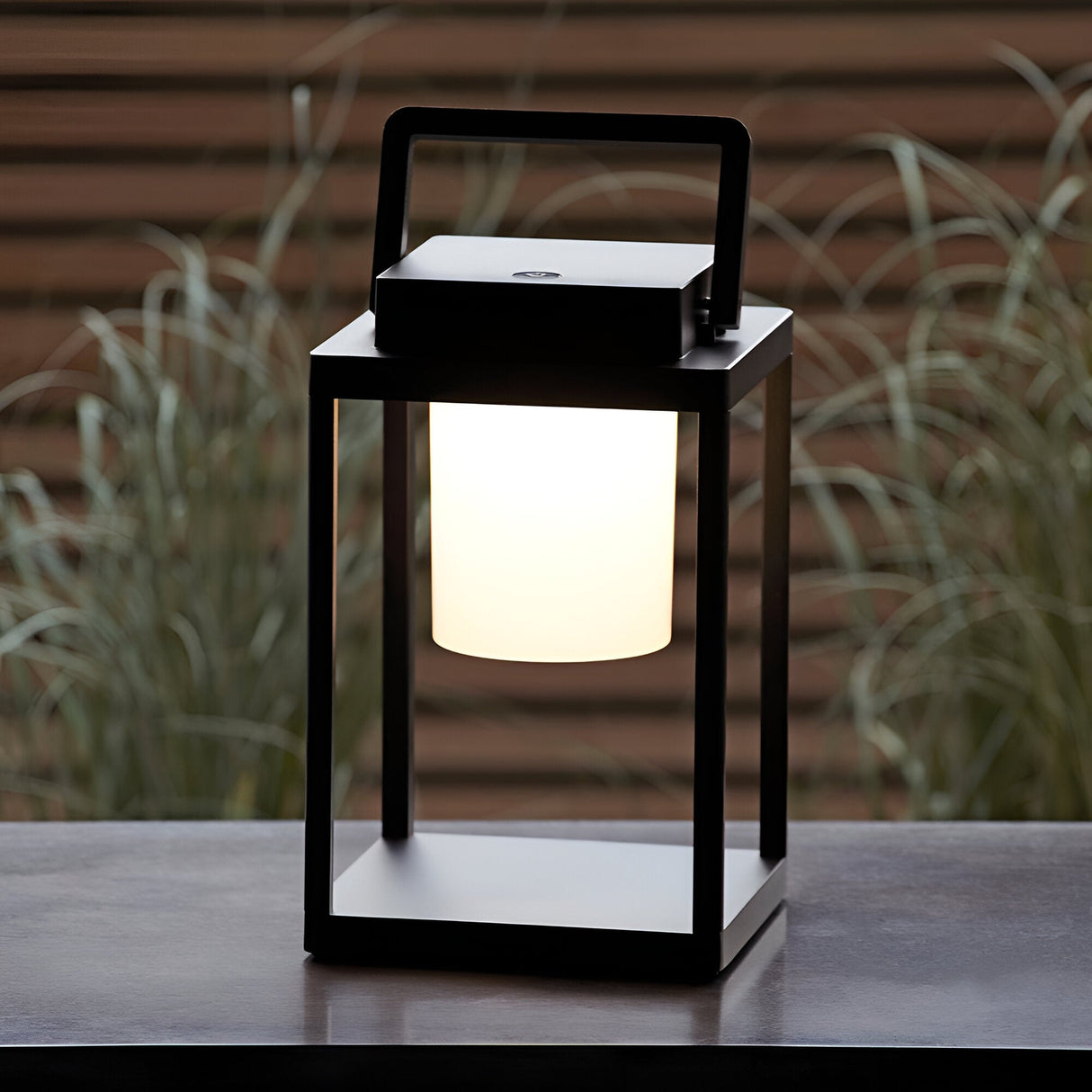 Endon Danedon portable LED table lamp in matt black with white diffuser and carry handle, on an outdoor table beside ornamental grasses.