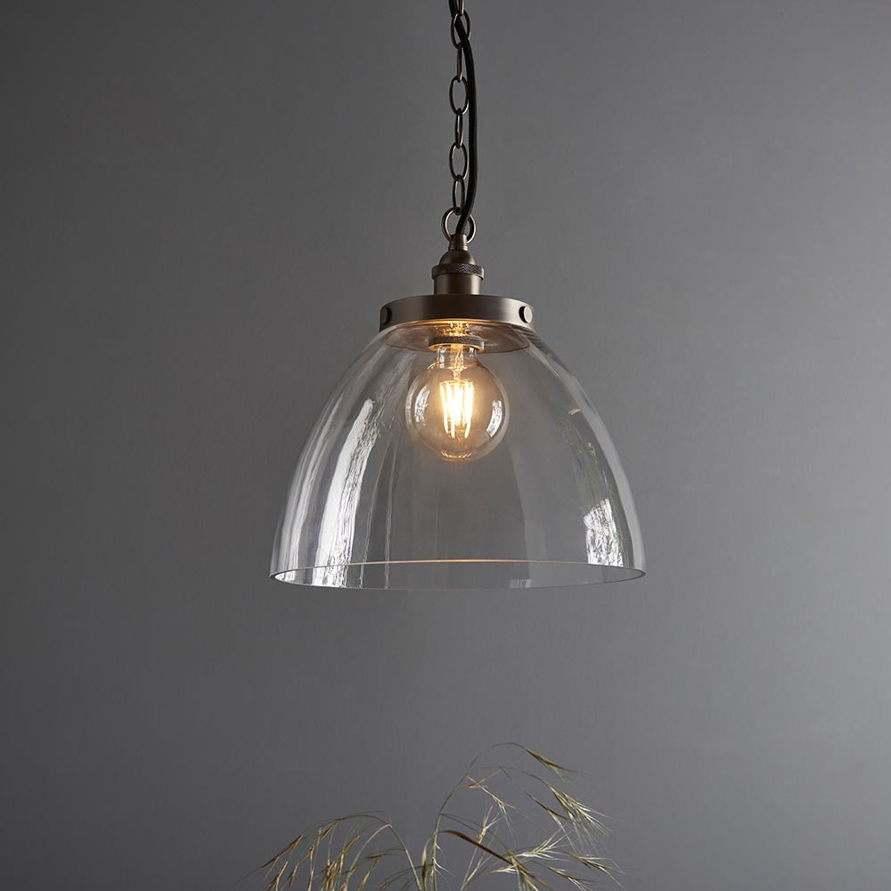 Wentworth Pendant in Silver, clear glass dome shade with brushed silver knurled lampholder on a black chain, filament bulb lit, against a grey background.