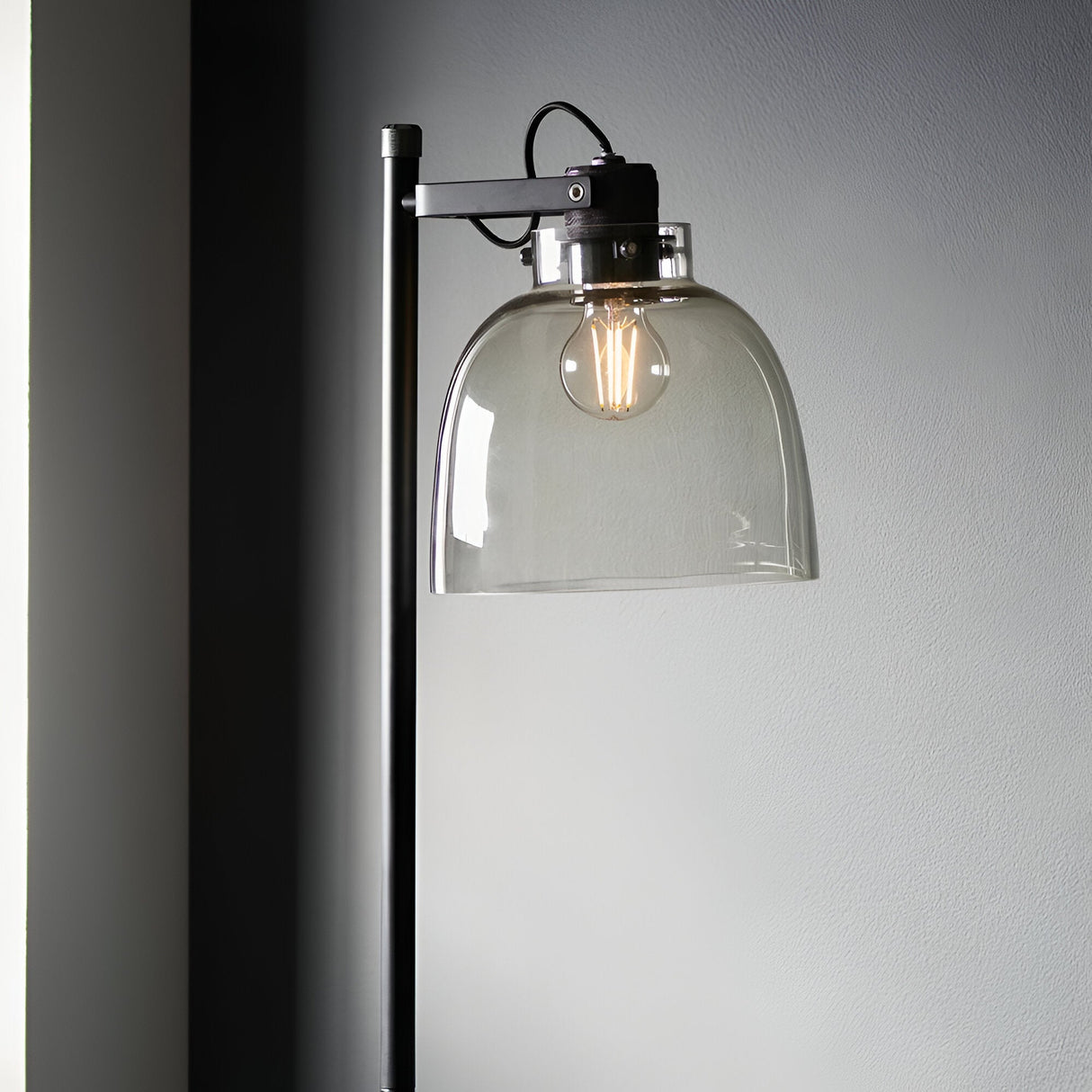 Endon Camden Task Floor Lamp in matt black with adjustable head, smoked grey glass shade and exposed filament bulb, set against a grey wall.