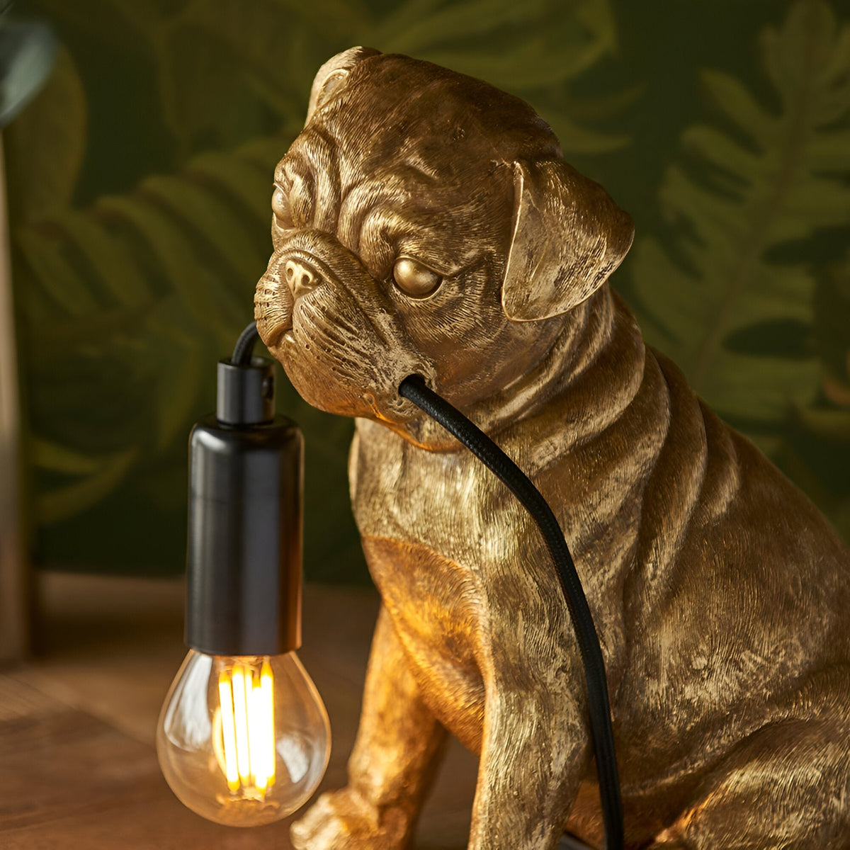 Endon Charthurst table lamp with a gold resin pug figure holding a black cable and exposed filament E14 bulb, set against leafy green decor.