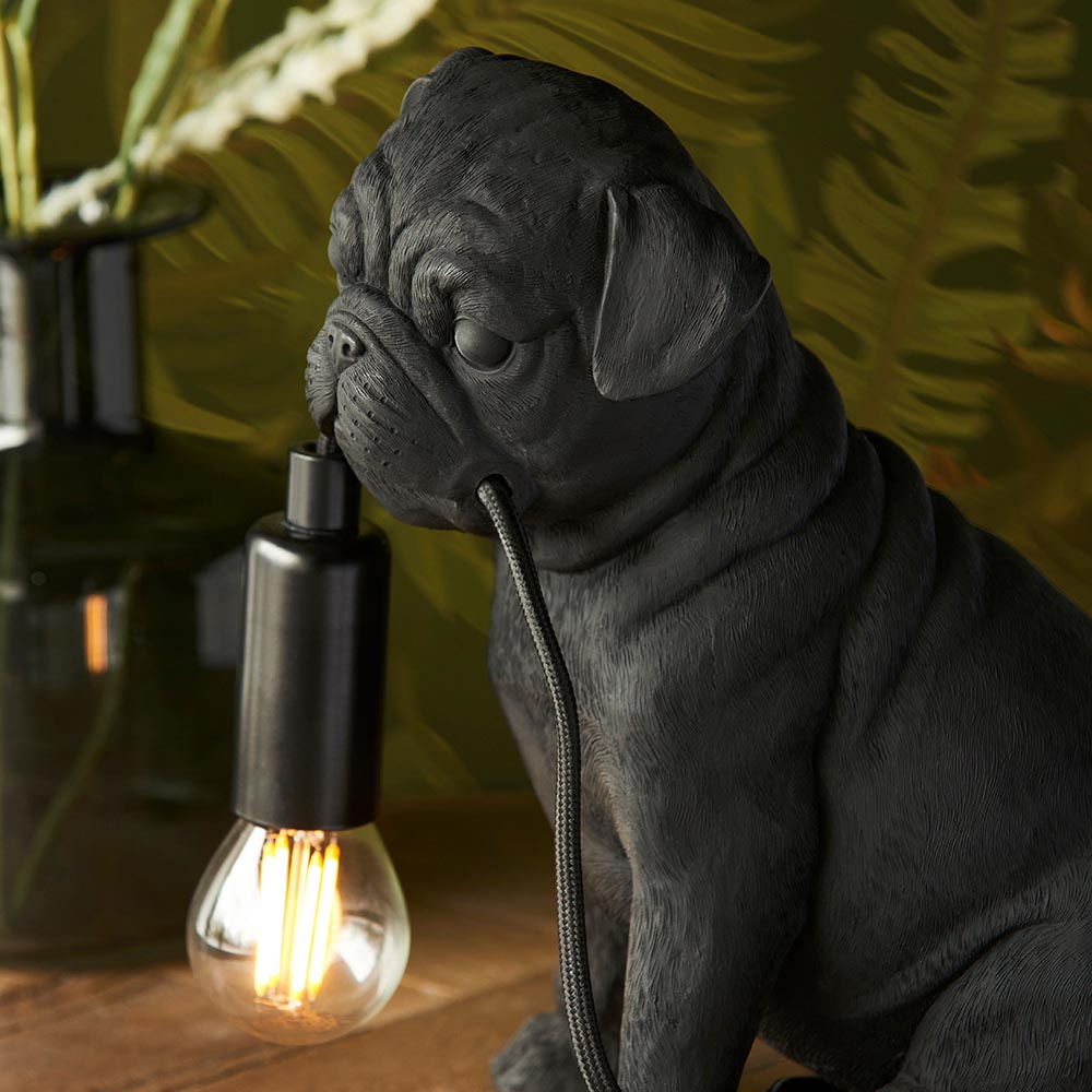 Endon Charthurst table lamp shaped as a matt black resin pug figure holding an E14 filament bulb holder, set on wood against a leafy backdrop.