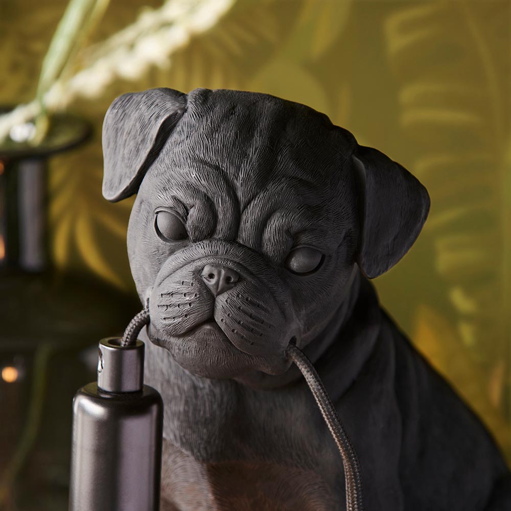 Endon Charthurst table lamp shaped as a detailed matt black resin pug figure, shown close-up with black fabric cable against a leafy green backdrop.