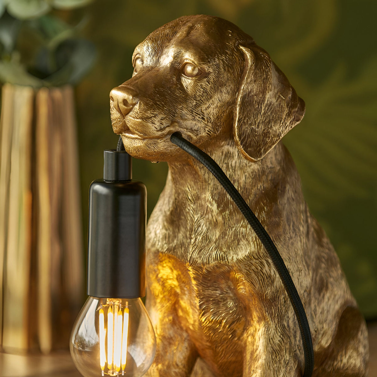 Brockworth Table Lamp in gold resin, shaped as a seated Labrador, with black lamp holder and filament bulb, set against a green background.