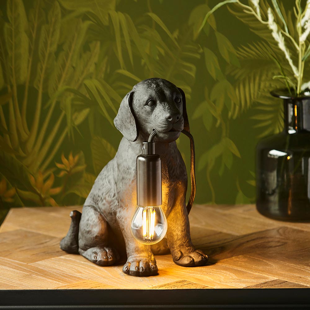 Endon Brockworth matt black resin Labrador puppy table lamp holding an E14 filament bulb, on a wooden surface against leafy green wallpaper.