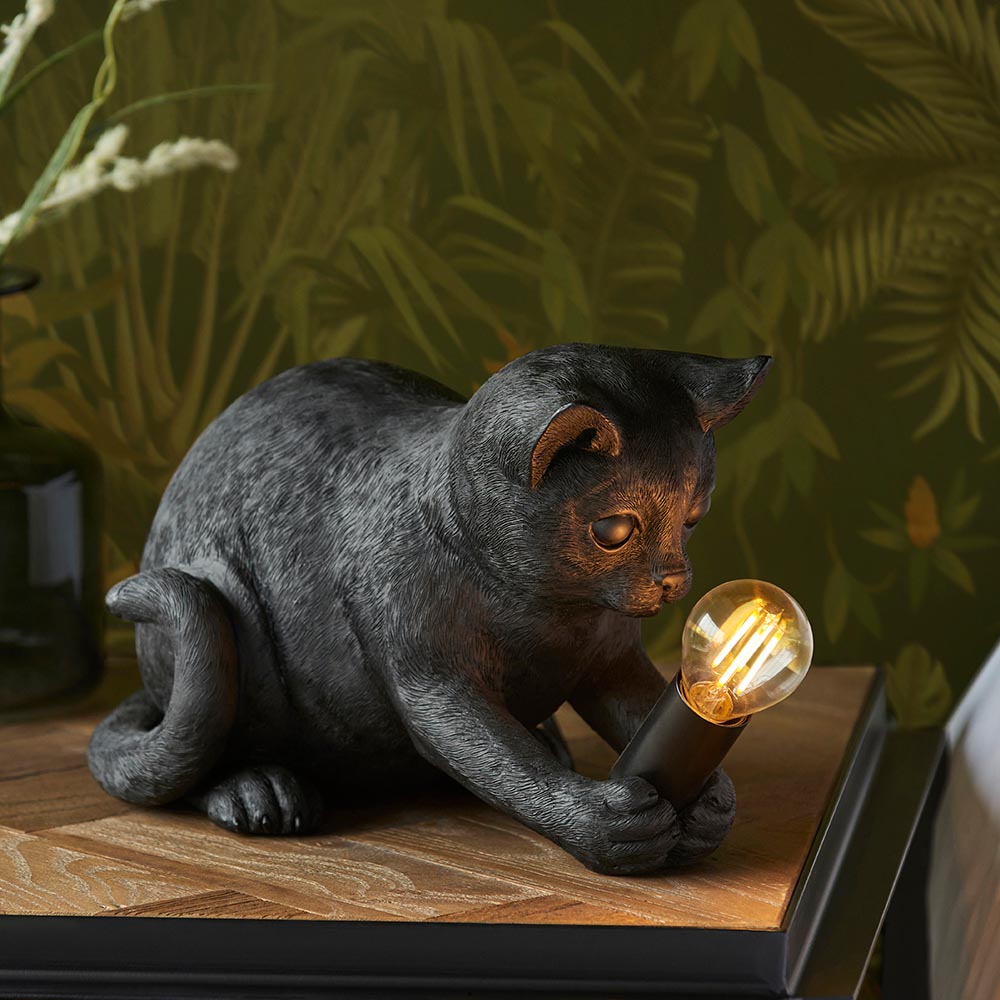 Endon Brockwick matt black resin kitten table lamp holding an E14 filament bulb, shown on a wooden side table against foliage wallpaper.