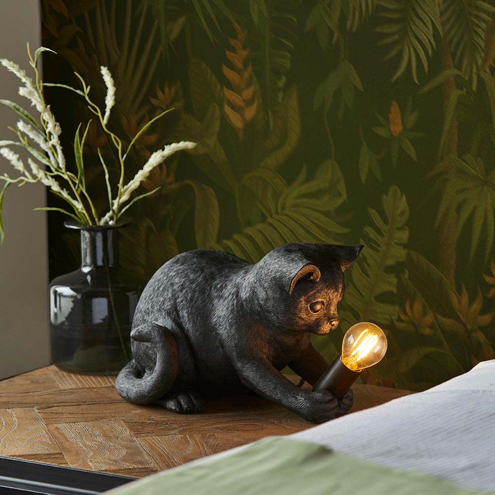 Endon Brockwick matt black resin kitten table lamp with visible filament E14 bulb, on a wooden bedside beside a smoked glass vase against botanical wallpaper.