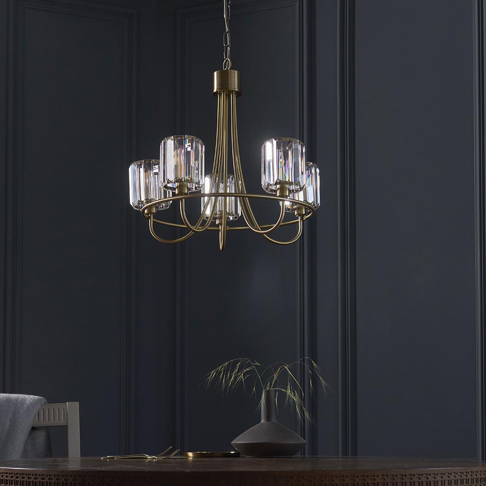 Endon Clementine 5 light pendant in brass with curved arms and cut faceted glass shades, hanging above a dining table against a dark panelled wall.