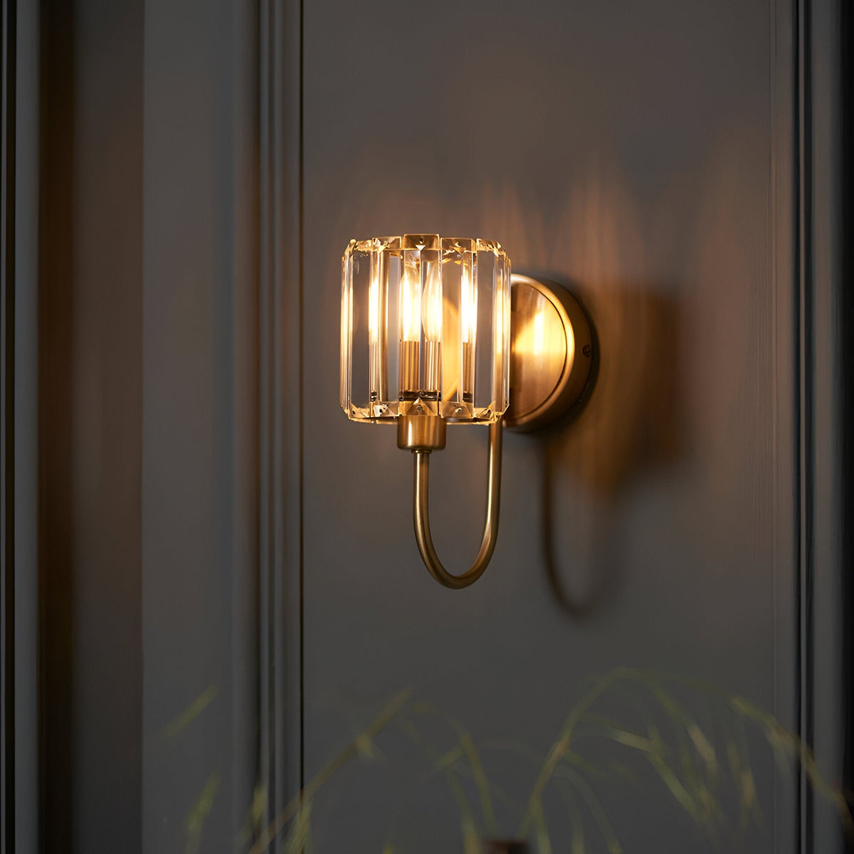 Endon Clementine wall light in brass with a cut faceted glass shade and curved arm, mounted on a grey panelled wall for ambient bedroom or hallway lighting.