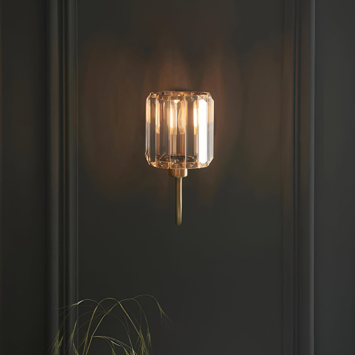 Endon Clementine wall light in brass with a cut faceted glass shade illuminated against a dark panelled wall.