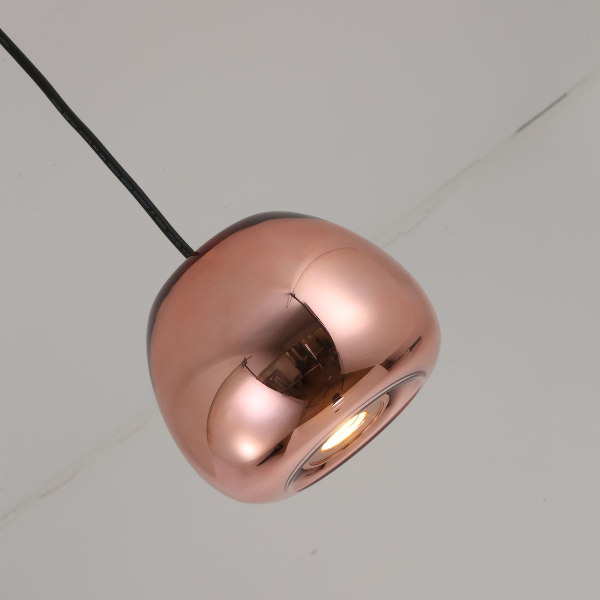 The Gulf Mini Glass Pendant Light - Copper features a shiny, metallic finish and hangs from a black cord against a plain white ceiling, casting a warm glow when illuminated.