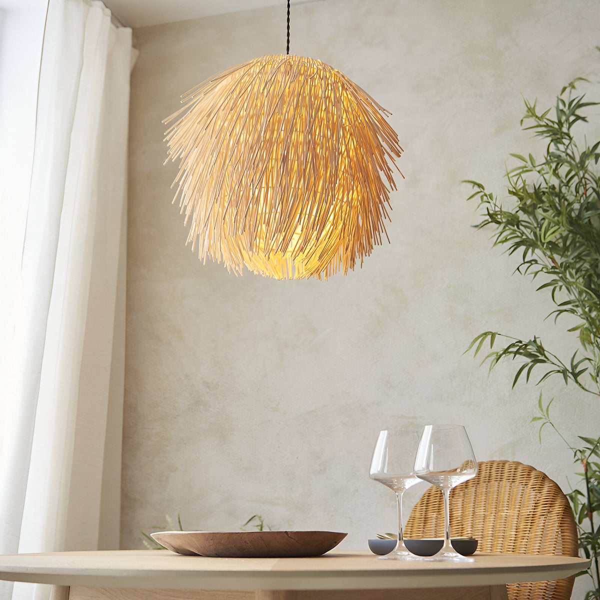 Chartleigh Pendant Shade in Natural rattan, a spiky layered globe design lit warmly above a dining table with wine glasses and a wicker chair.