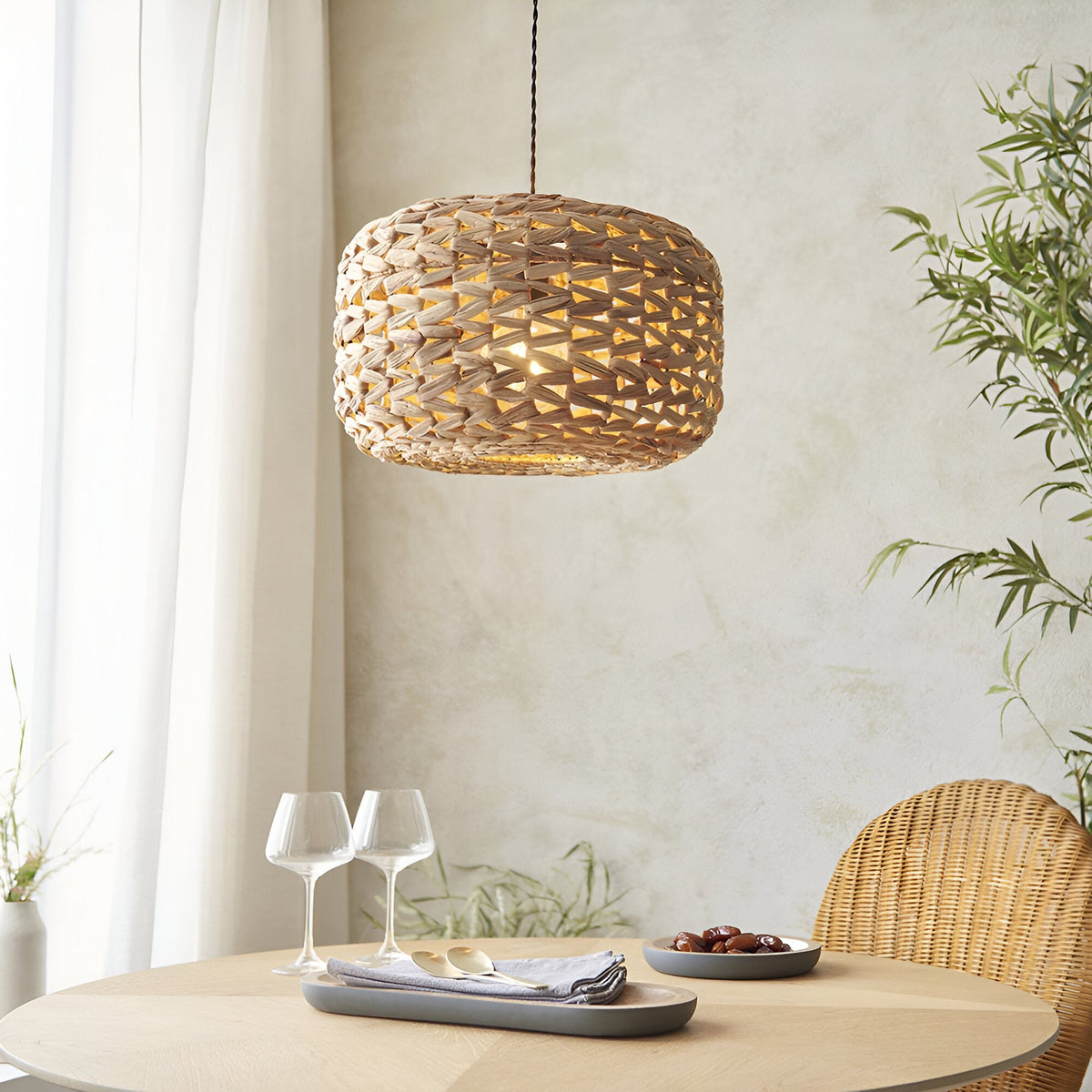 Endon Cranley natural handwoven rattan pendant shade with a soft curved drum shape, illuminated above a dining table beside a wicker chair and bamboo plant.