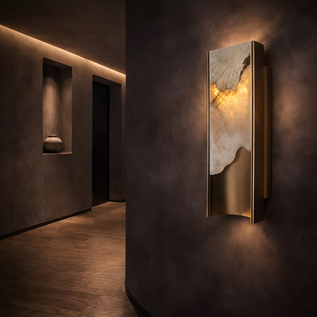 A modern hallway with dim lighting features the Veloris LED Small Carved Alabaster Wall Light in brushed brass & soft white, providing a warm accent. A niche with a ceramic vase and an open doorway are visible in the background.