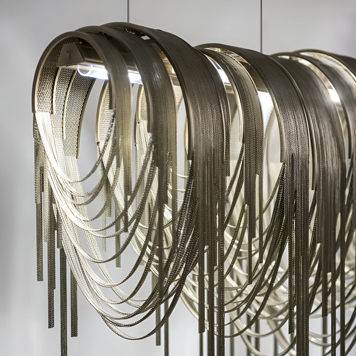A close-up of the Symphonia 10 Light Metal Chain Veil Suspension in Gold, featuring curved metallic strips and hanging chains, illuminated from above for a modern, elegant look against a plain background.