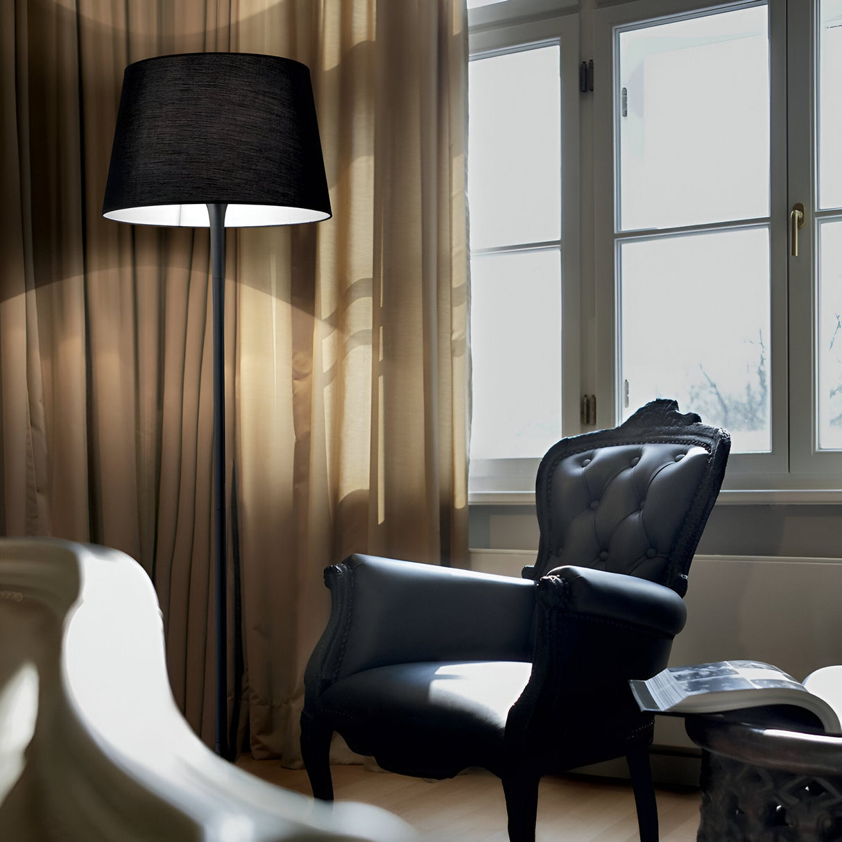 In the cozy living room, minimalist elegance is showcased with a black leather armchair and an Arc Floor Lamp - Black, casting a warm glow. Sheer curtains gently cover the window, enhancing the soft ambiance, while a coffee table with a curved design is partially visible.