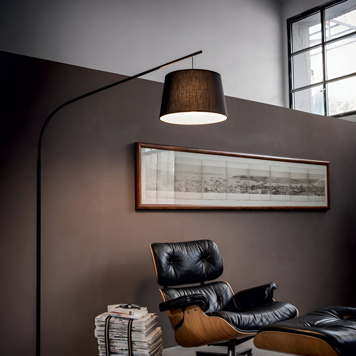 A modern living room features a black leather Eames lounge chair, illuminated by the Luna Floor Lamp - Black, which provides elegant ambient lighting. A stack of magazines rests nearby, beneath a large window and framed landscape artwork that complement the brown wall.