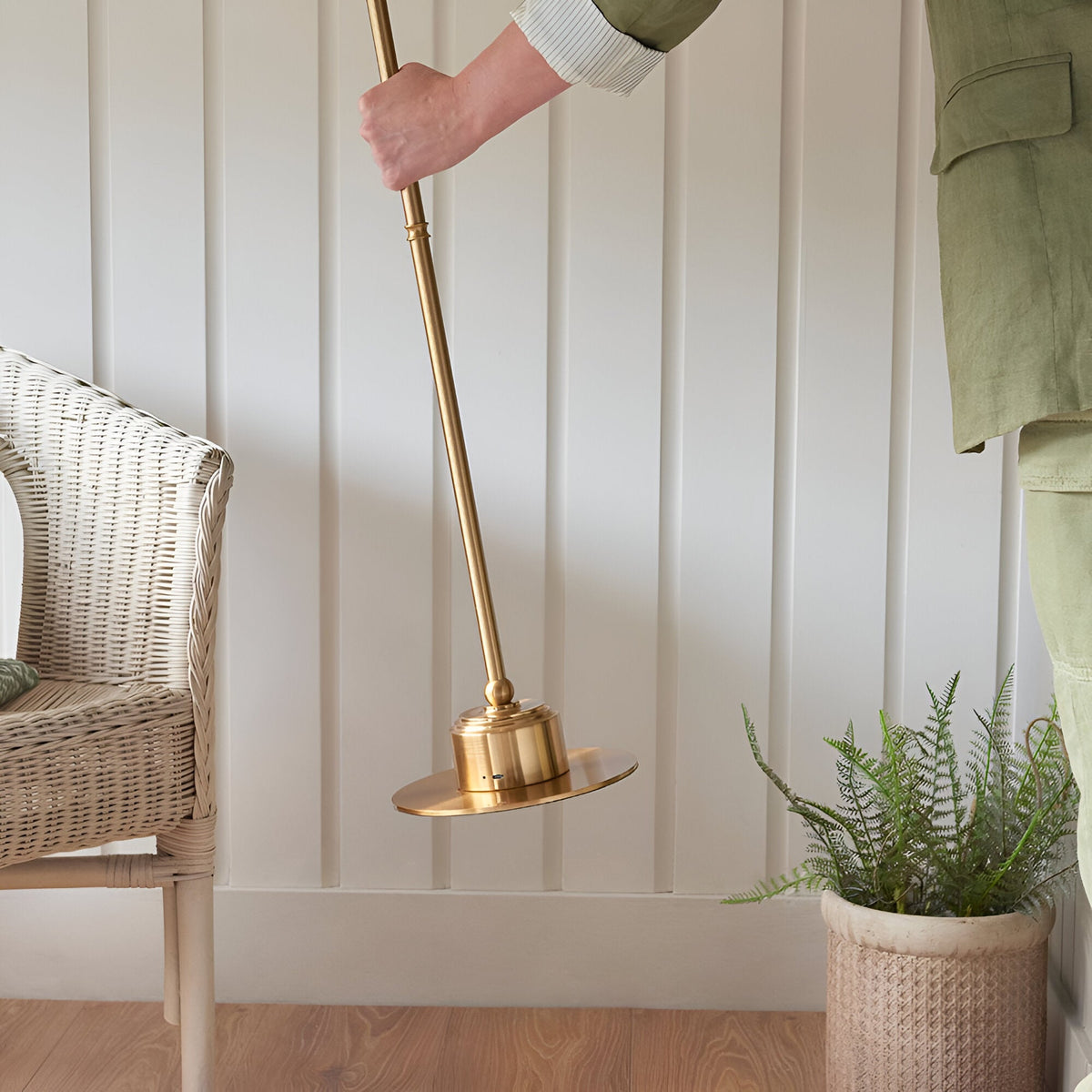 Hand lifting the Endon Ashstead rechargeable floor lamp by its slim aged brass stem, showing the round base beside a wicker chair and potted fern.