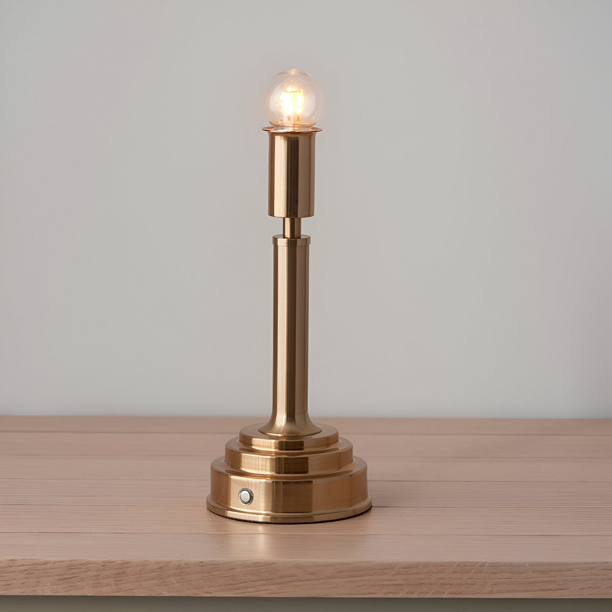 Danebourne LED Table Lamp in brushed aged brass with stepped base and exposed globe bulb, shown lit on a wood surface against a plain grey background.