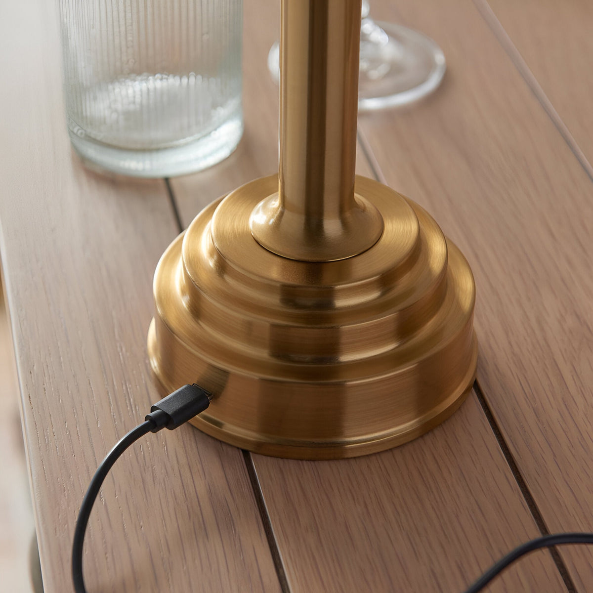 Danebourne LED Table Lamp in brushed antique brass, stepped tiered base with USB charging cable, shown close-up on a wooden table.