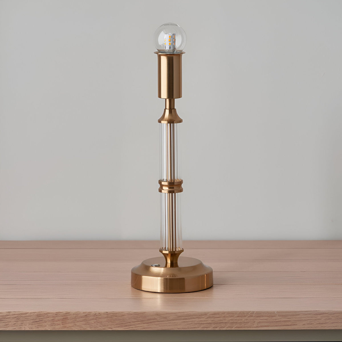 Endon Carlwood LED table lamp in brushed aged brass with clear acrylic column detail and exposed bulb, shown on a light wood surface.