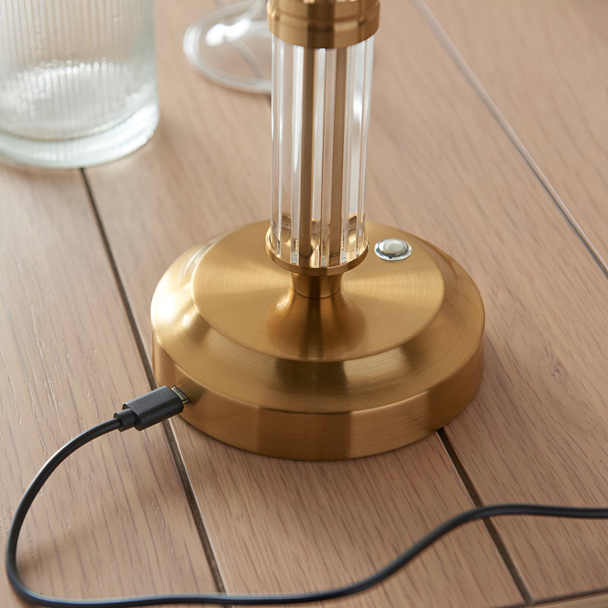 Close-up of Endon Carlwood LED table lamp brass base with clear acrylic detail, touch dimmer button and USB-C charging cable connected, on wooden surface.