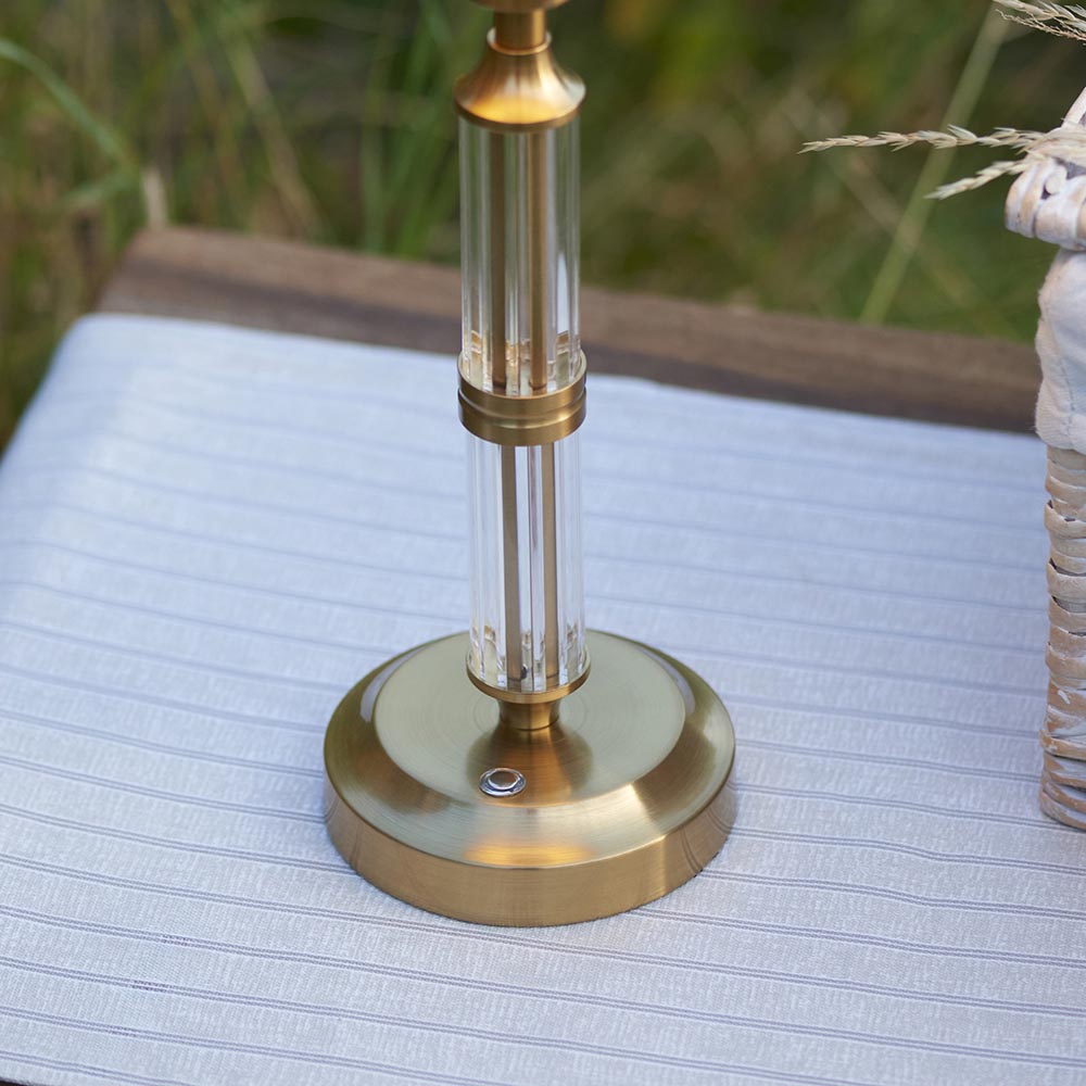 Endon Carlwood LED table lamp base in brushed aged brass with clear acrylic detailing and touch dimmer button, set on a striped cushion outdoors.