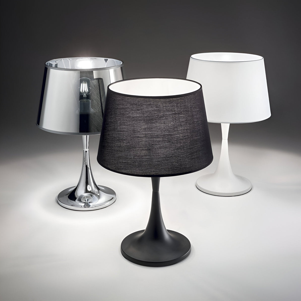 On display are three table lamps, including the Arc Small Table Lamp - White. It features a minimalist design and sleek curves, perfect for modern lighting in compact spaces, and stands elegantly on a reflective surface; accompanied by options with metallic silver and black shades.