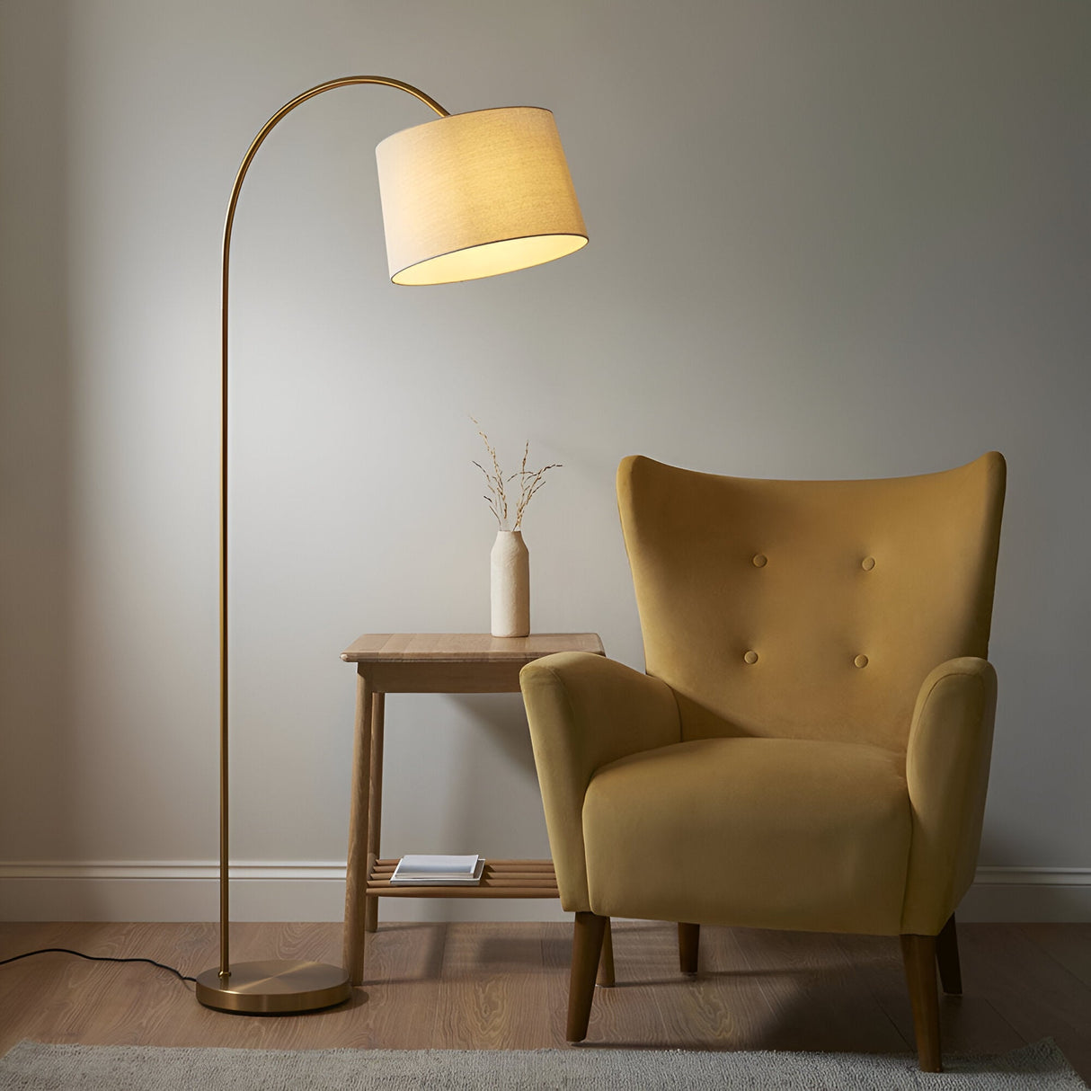 Endon Bevbourne arched floor lamp in brass with tilting natural fabric drum shade, beside a mustard velvet armchair and wooden side table.