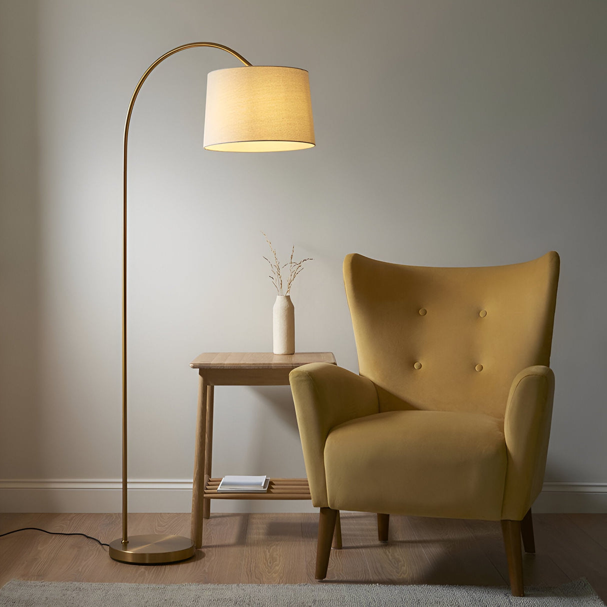 Endon Bevbourne arched floor lamp in brass with tilting natural fabric drum shade, beside a mustard velvet armchair and wooden side table.
